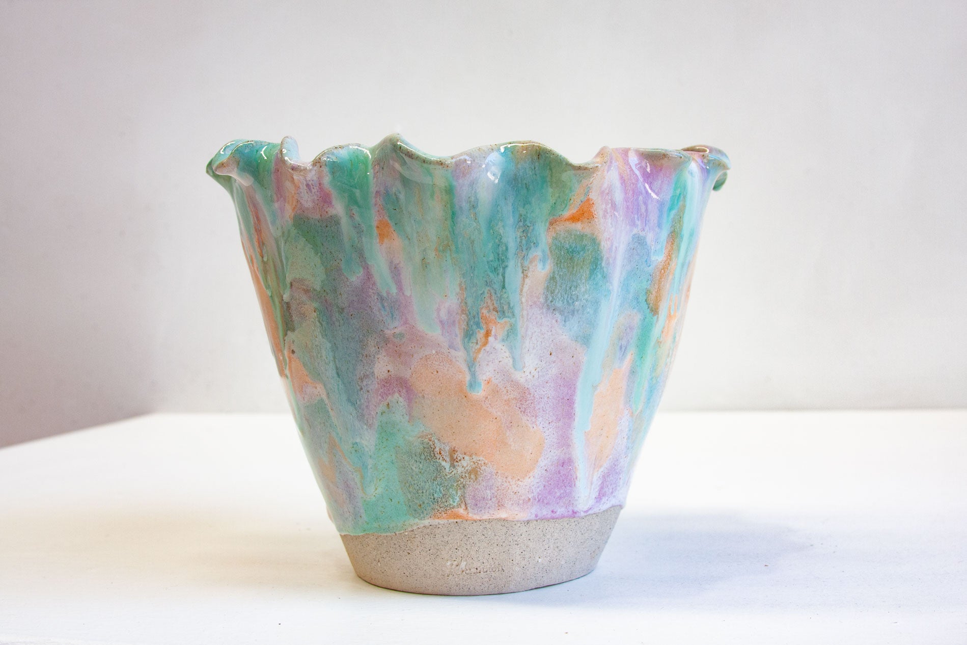 A handmade ceramic frill vessel seen from the side, the frilled rim catching the light above soft bands of purple, peach and green.