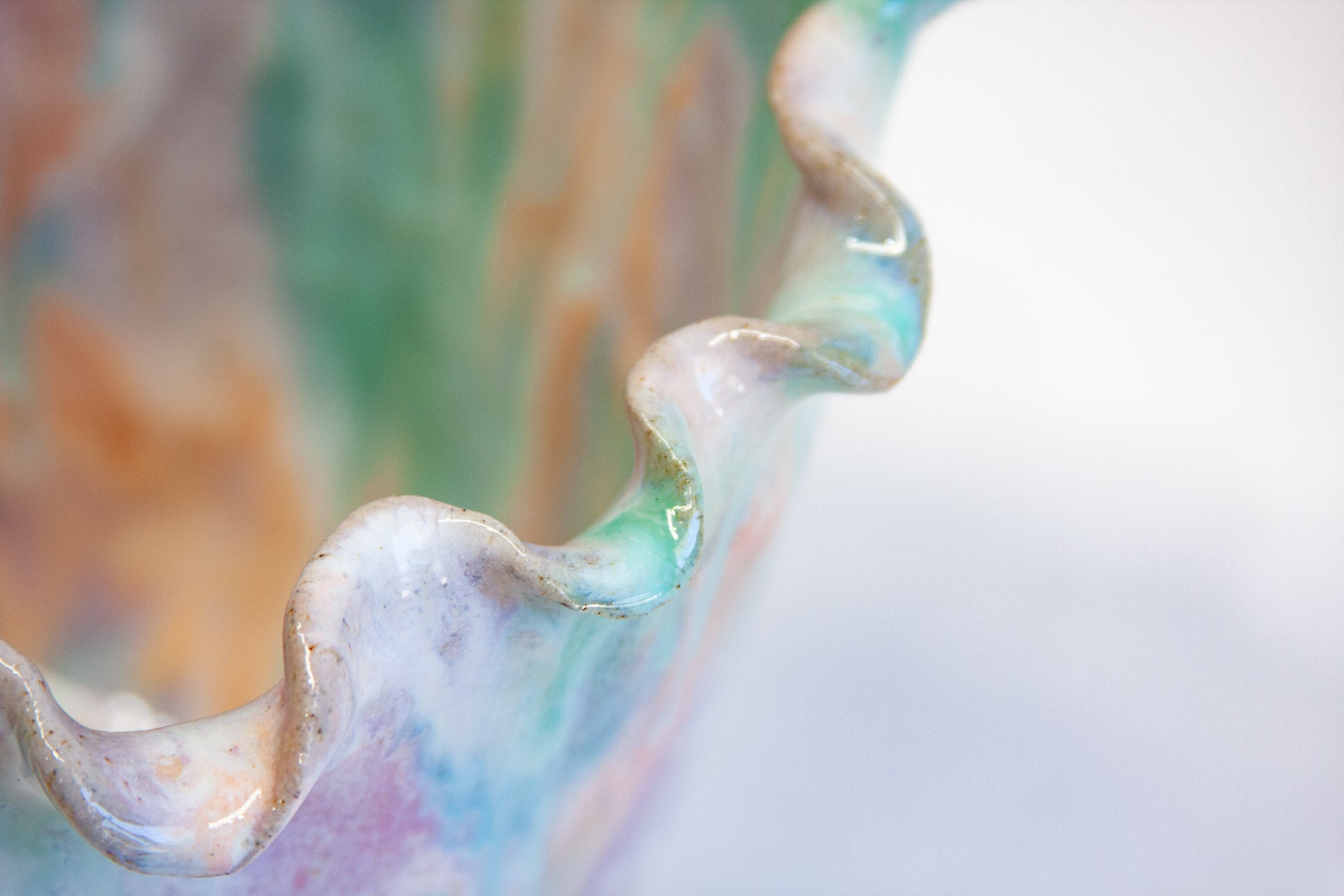 Close-up of the frilled rim of a handmade ceramic frill vessel, pale green and blue along the folded edge above streaks of purple and peach.