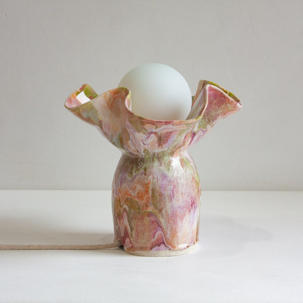 Front-on view of lit handmade ceramic table lamp, bell-shaped base with wide ruffled petal top glazed in swirling pink, mauve, peach, amber and sage green, glowing white globe bulb, on a white surface