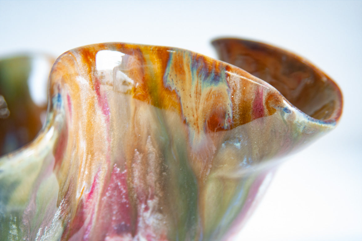 Extreme close-up of the ruffled rim edge of the dark clay oyster bowl; streaked glaze in amber-orange, deep pink, teal, and pale olive visible in fine detail.