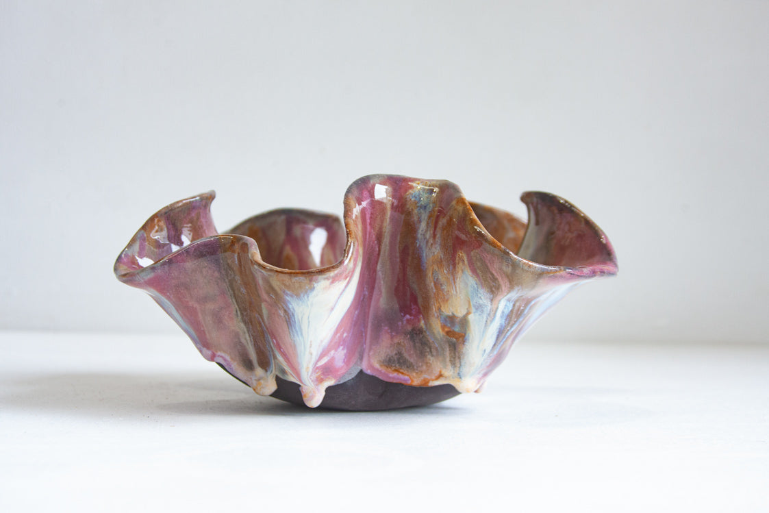 Low front angle of handmade dark clay oyster bowl showing the unglazed dark base and tall ruffled walls; glaze in dusky pink, amber, and pale blue-white drapes down the exterior flanges.