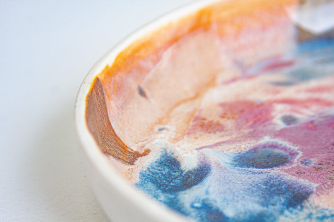 Close-up angled view of the interior rim of a handmade ceramic deep plate showing amber, brown, pink and blue paint-pour glaze