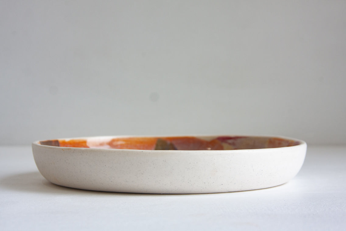 Side profile view of a handmade ceramic deep plate showing unglazed white speckled stoneware exterior, amber and orange paint-pour glaze visible at interior rim