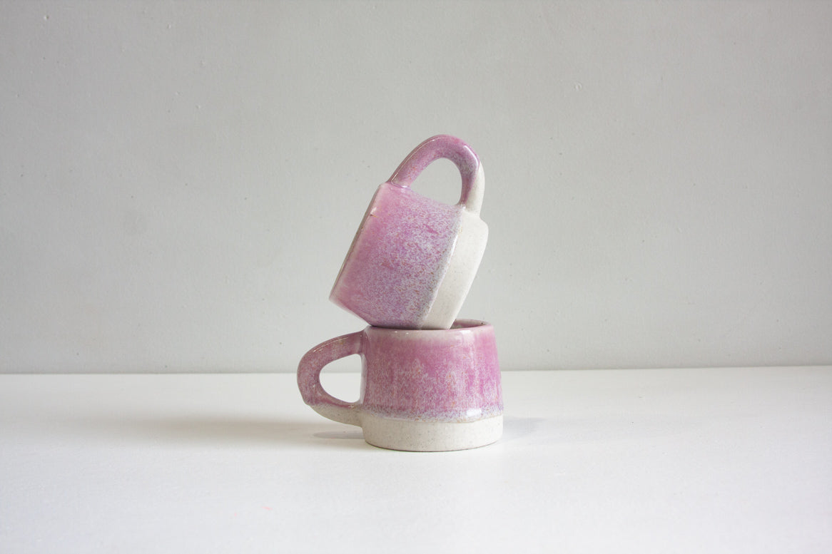 Two small ceramic mugs with a pink and white speckled glaze, stacked with one tilted on top of the other, on a white surface against a light gray background.