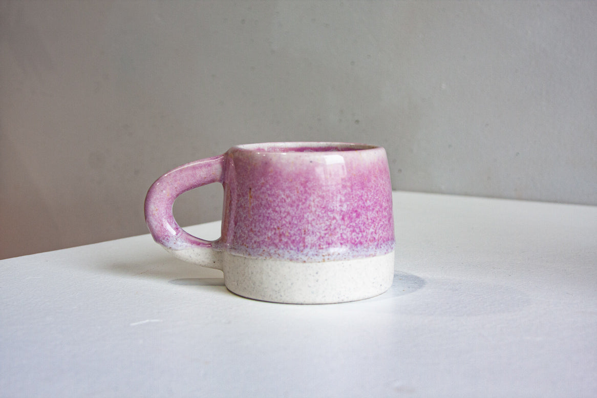 A handmade ceramic mug with a pink speckled glaze on the upper portion and a textured off-white unglazed base, sitting on a white surface against a neutral gray background.