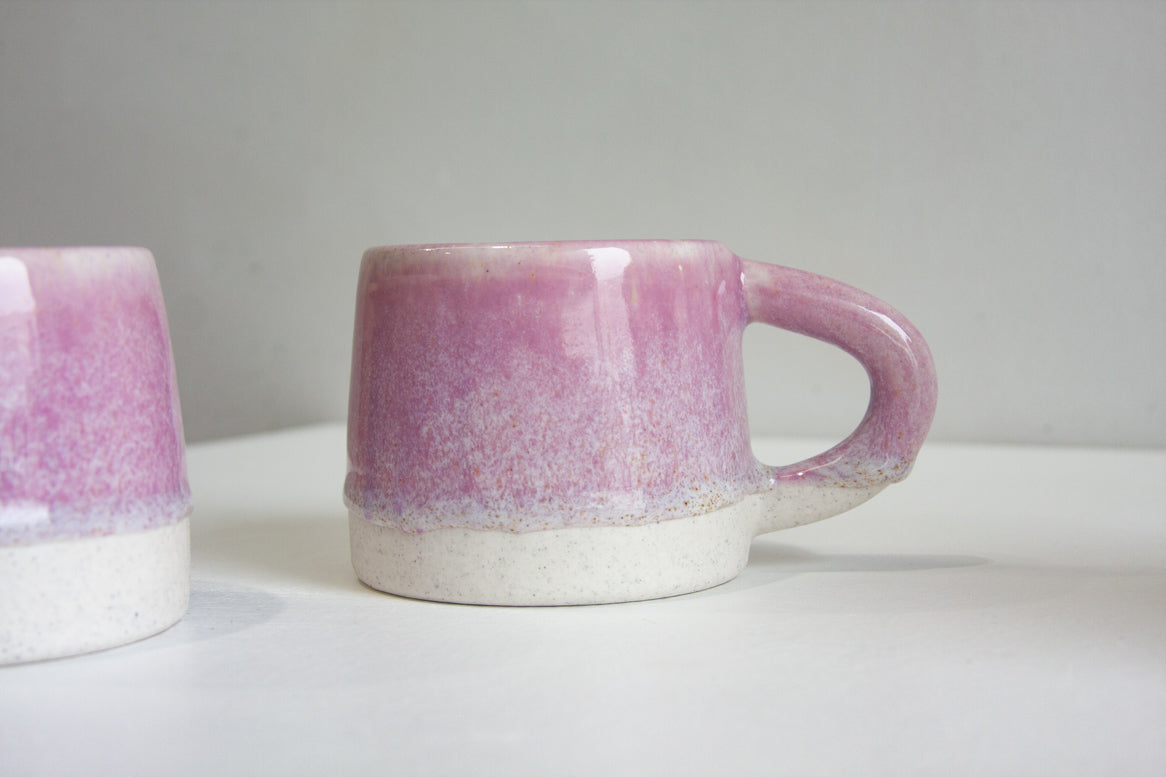 A handmade ceramic mug with a pink speckled glaze and a white unglazed base, shown on a white surface against a gray background.
