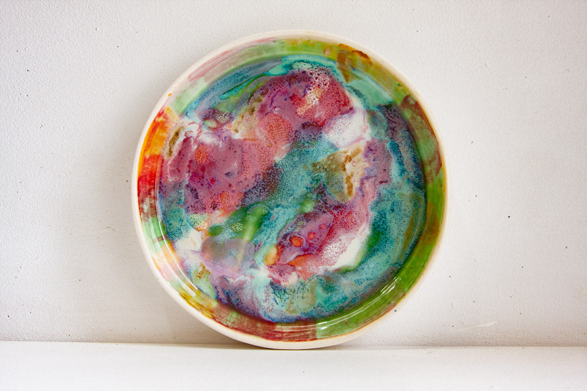 Front-on view of a handmade ceramic deep plate propped upright, vivid multicolour paint-pour glaze in pink, teal, green, red, orange and amber on white stoneware