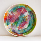 Front-on view of a handmade ceramic deep plate propped upright, vivid multicolour paint-pour glaze in pink, teal, green, red, orange and amber on white stoneware