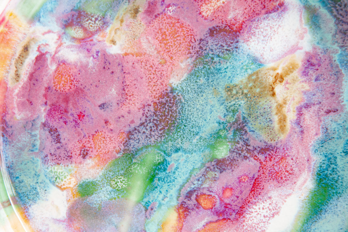Extreme close-up of the interior glaze surface of a handmade ceramic deep plate showing swirling pink, teal, green, orange and amber paint-pour detail