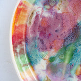 Close-up angled view of the interior of a handmade ceramic deep plate showing vivid pink, purple, teal, green, orange and amber paint-pour glaze with unglazed rim visible