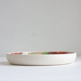Side profile view of a handmade ceramic deep plate showing the low wide form and unglazed white stoneware exterior, multicolour glaze just visible at the interior rim