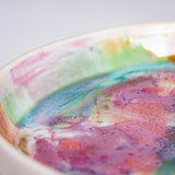 Close-up angled view of the interior rim of a handmade ceramic deep plate showing multicolour paint-pour glaze detail in pink, teal, green and orange