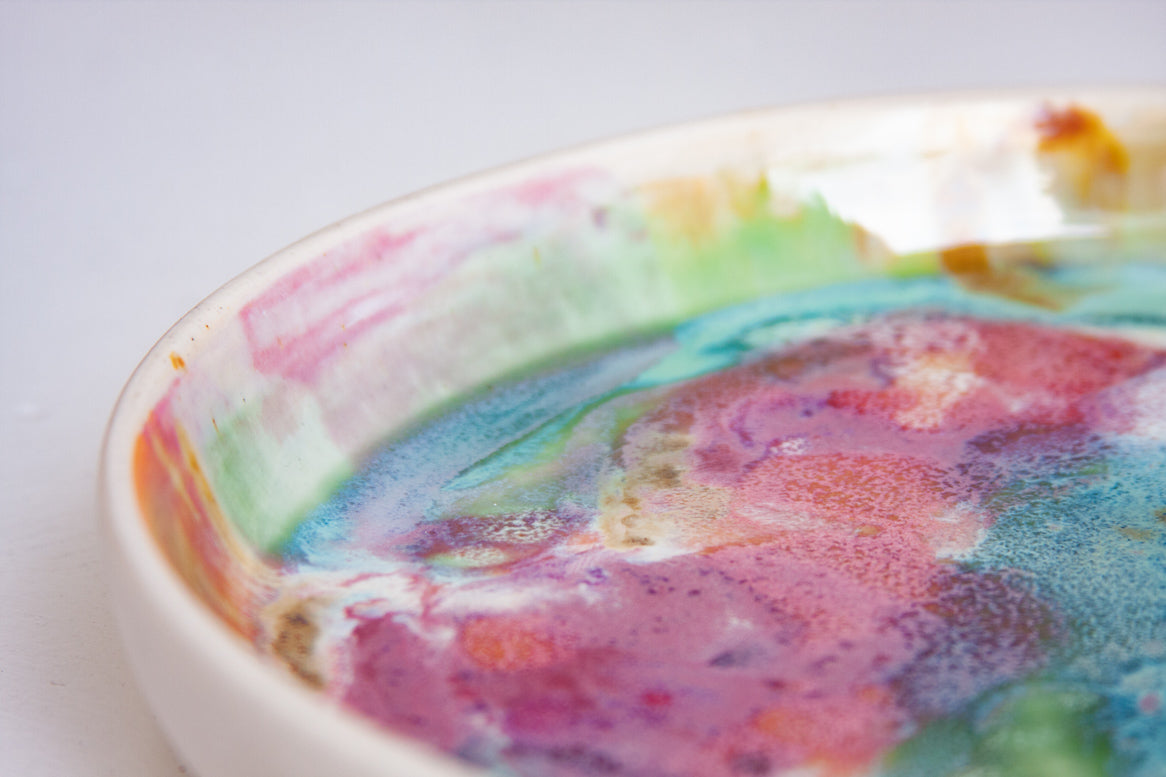 Close-up angled view of the interior rim of a handmade ceramic deep plate showing multicolour paint-pour glaze detail in pink, teal, green and orange