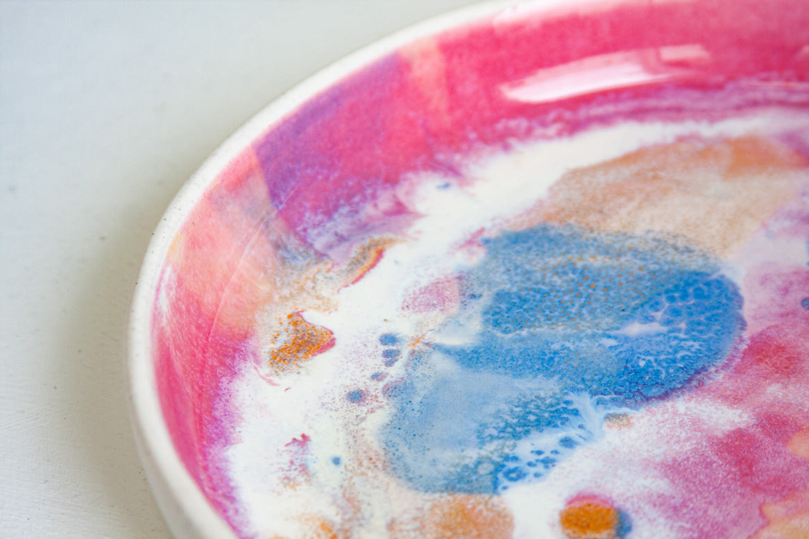 Close-up angled view of the interior rim of a handmade ceramic deep plate with vibrant pink and purple rim and swirling blue, orange and white paint-pour glaze