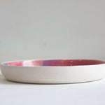 Side profile view of a handmade ceramic deep plate with unglazed white stoneware exterior, pink and blue glaze just visible at the interior rim