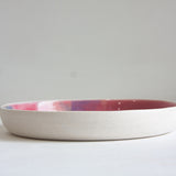 Side profile view of a handmade ceramic deep plate with unglazed white stoneware exterior, pink and blue glaze just visible at the interior rim