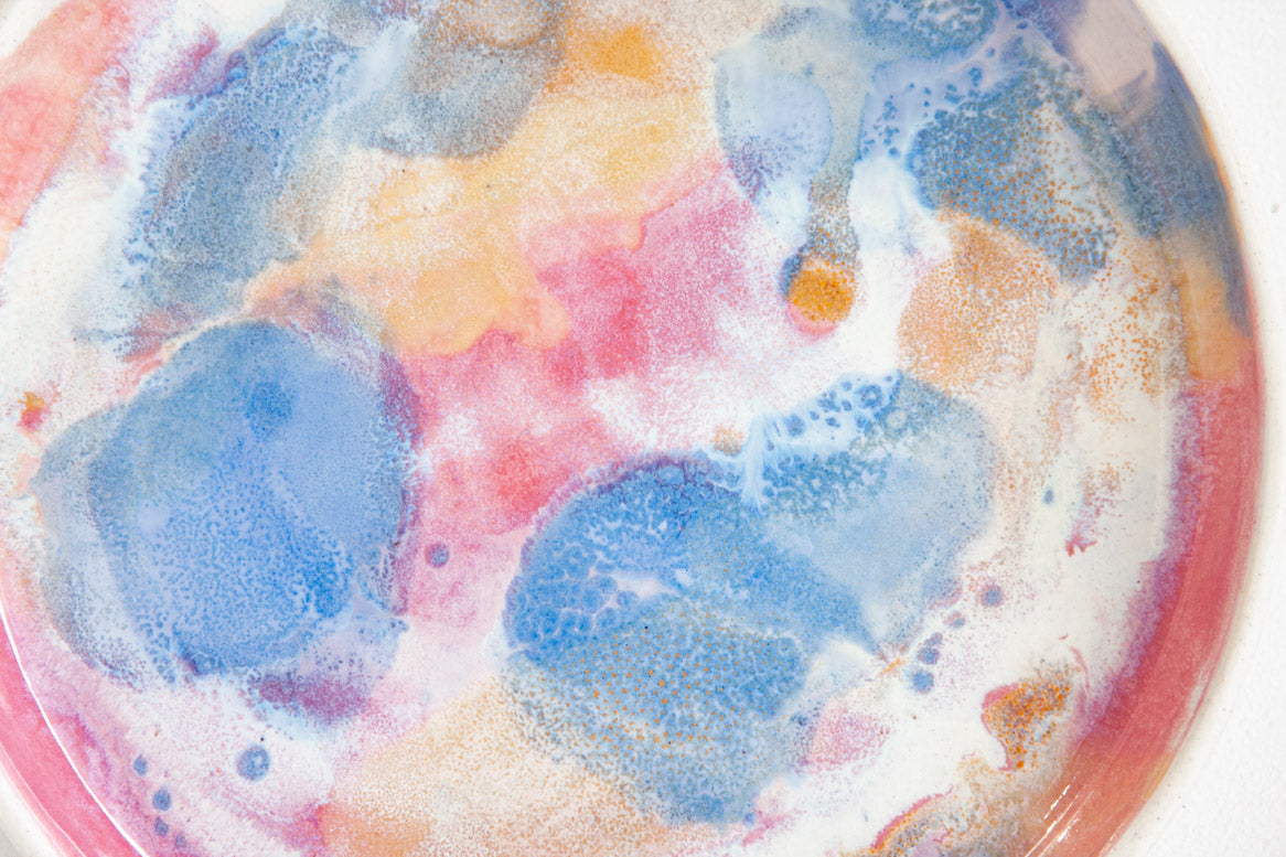 Top-down interior view of a handmade ceramic deep plate with soft blue, pink, peach and amber paint-pour glaze on a white base