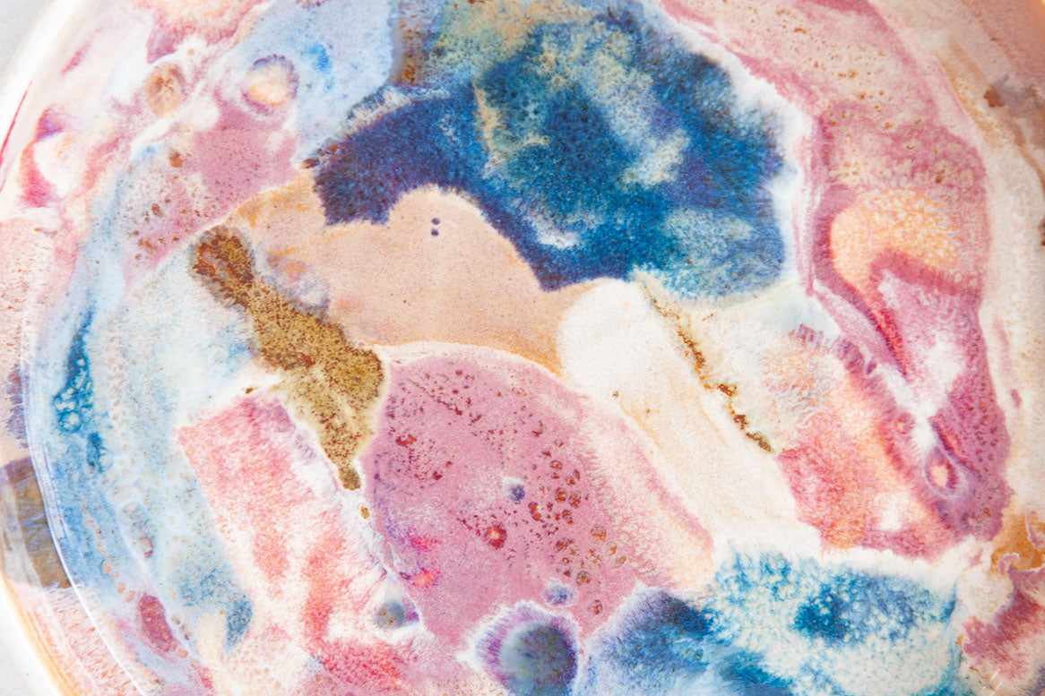 Extreme close-up of the interior of a handmade ceramic deep plate showing swirling blue, pink, mauve, amber and white paint-pour glaze detail