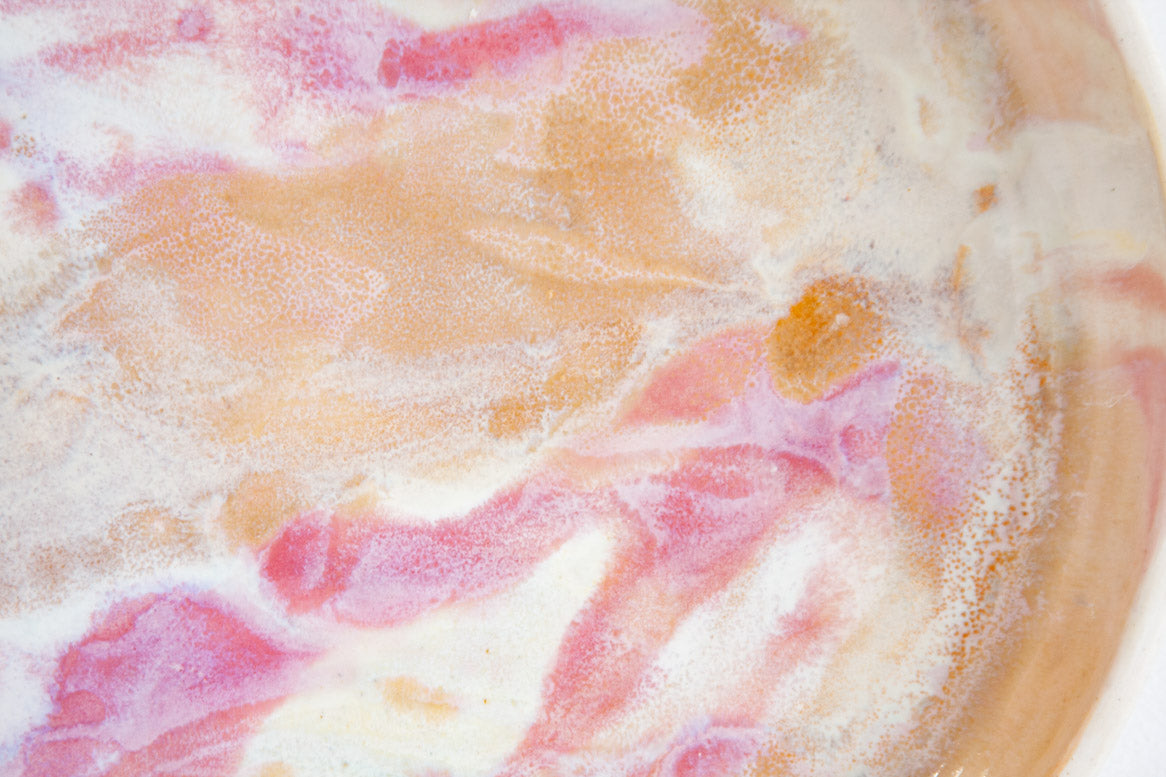 Extreme close-up of the interior of a handmade ceramic deep plate showing swirling soft pink, peach, amber and white paint-pour glaze detail