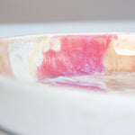 Close-up side view of the interior rim of a handmade ceramic deep plate with soft pink, peach and amber paint-pour glaze on white stoneware