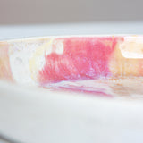 Close-up side view of the interior rim of a handmade ceramic deep plate with soft pink, peach and amber paint-pour glaze on white stoneware