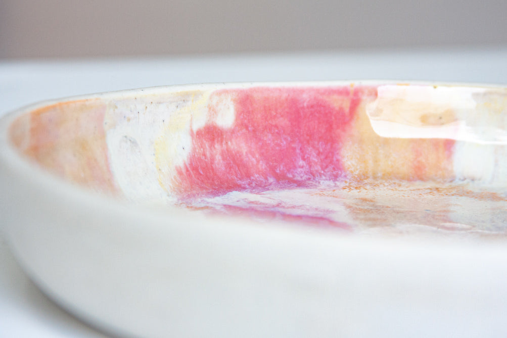 Close-up side view of the interior rim of a handmade ceramic deep plate with soft pink, peach and amber paint-pour glaze on white stoneware