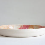 Side profile view of a handmade ceramic deep plate showing unglazed white speckled stoneware exterior, soft pink, peach and amber glaze visible at interior rim
