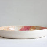 Side profile view of a handmade ceramic deep plate showing unglazed white speckled stoneware exterior, soft pink, peach and amber glaze visible at interior rim