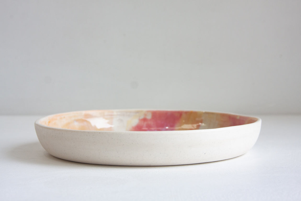 Side profile view of a handmade ceramic deep plate showing unglazed white speckled stoneware exterior, soft pink, peach and amber glaze visible at interior rim