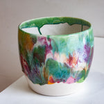 Three-quarter angled view of a handmade ceramic large planter showing green glazed interior and vivid multicolour drip glaze exterior on a white shelf