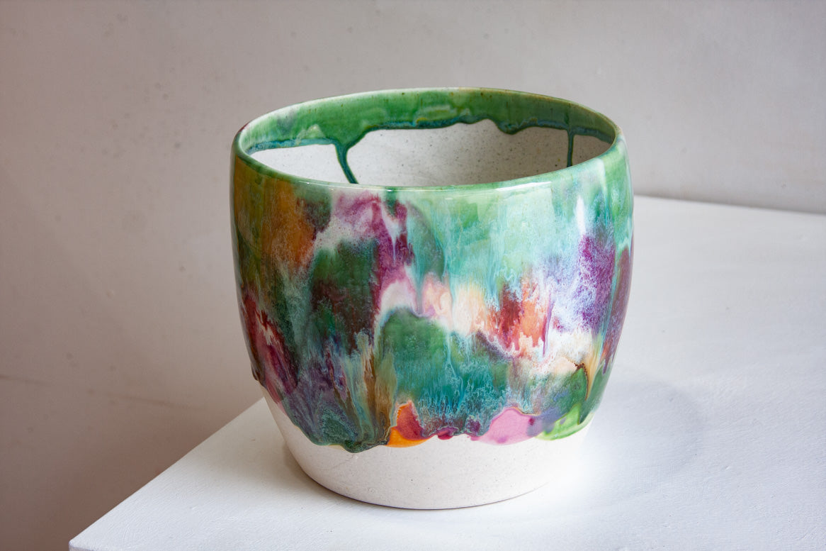 Three-quarter angled view of a handmade ceramic large planter showing green glazed interior and vivid multicolour drip glaze exterior on a white shelf