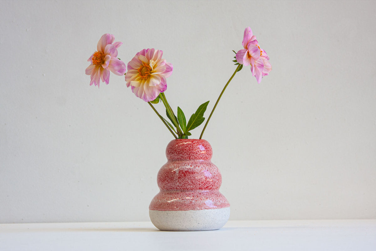 Lifestyle shot of handmade ceramic small smoosh vase with three stacked rounded lobes in speckled red-pink glaze over white speckled stoneware base, filled with pink dahlias