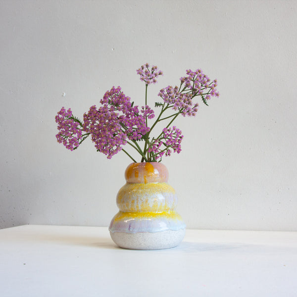 Lifestyle shot of handmade ceramic small smoosh vase with three stacked lobes, peach-pink and yellow drip glaze over soft lavender-white, unglazed speckled stoneware base, filled with pink yarrow, front-on view
