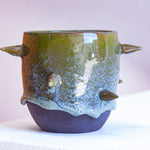 Front-facing side view of handmade dark clay spiked vessel against a pink background; cylindrical form with olive green drip glaze fading to speckled blue-grey; protruding pointed clay spikes; unglazed dark clay base.