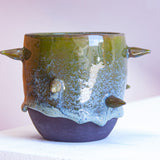 Front-facing side view of handmade dark clay spiked vessel against a pink background; cylindrical form with olive green drip glaze fading to speckled blue-grey; protruding pointed clay spikes; unglazed dark clay base.