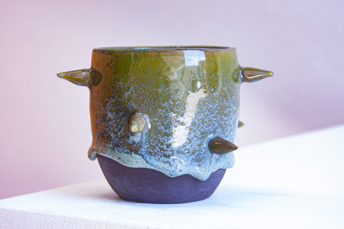 Front-facing side view of handmade dark clay spiked vessel against a pink background; cylindrical form with olive green drip glaze fading to speckled blue-grey; protruding pointed clay spikes; unglazed dark clay base.