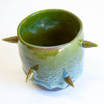Three-quarter overhead view of empty handmade dark clay spiked vessel; cylindrical form with deep olive green glaze fading to pale speckled blue-green; six protruding pointed clay spikes; dark unglazed base.