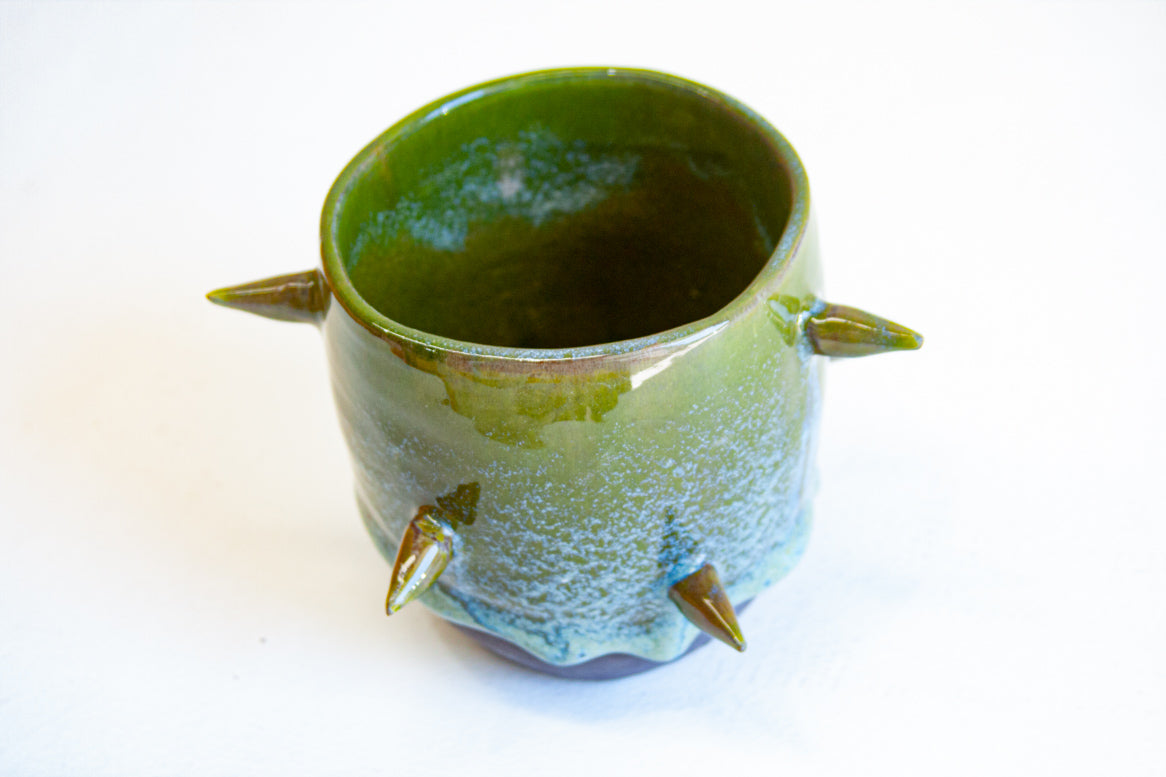Three-quarter overhead view of empty handmade dark clay spiked vessel; cylindrical form with deep olive green glaze fading to pale speckled blue-green; six protruding pointed clay spikes; dark unglazed base.