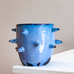 Front-facing side view of empty handmade dark clay spiked vessel on a white plinth against a pink background; cobalt-blue glaze with darker tones at the rim; protruding dark-tipped pointed spikes; unglazed dark clay base patches.
