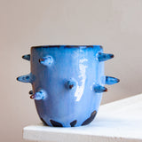 Front-facing side view of empty handmade dark clay spiked vessel on a white plinth against a pink background; cobalt-blue glaze with darker tones at the rim; protruding dark-tipped pointed spikes; unglazed dark clay base patches.
