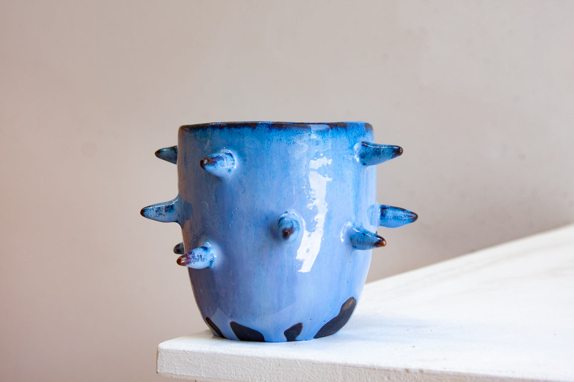 Front-facing side view of empty handmade dark clay spiked vessel on a white plinth against a pink background; cobalt-blue glaze with darker tones at the rim; protruding dark-tipped pointed spikes; unglazed dark clay base patches.
