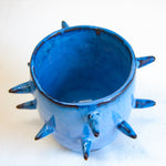 Three-quarter overhead view of empty handmade dark clay spiked vessel; rich cobalt-blue glaze covering the interior and exterior; multiple dark-tipped pointed spikes; dark unglazed clay visible at the base.