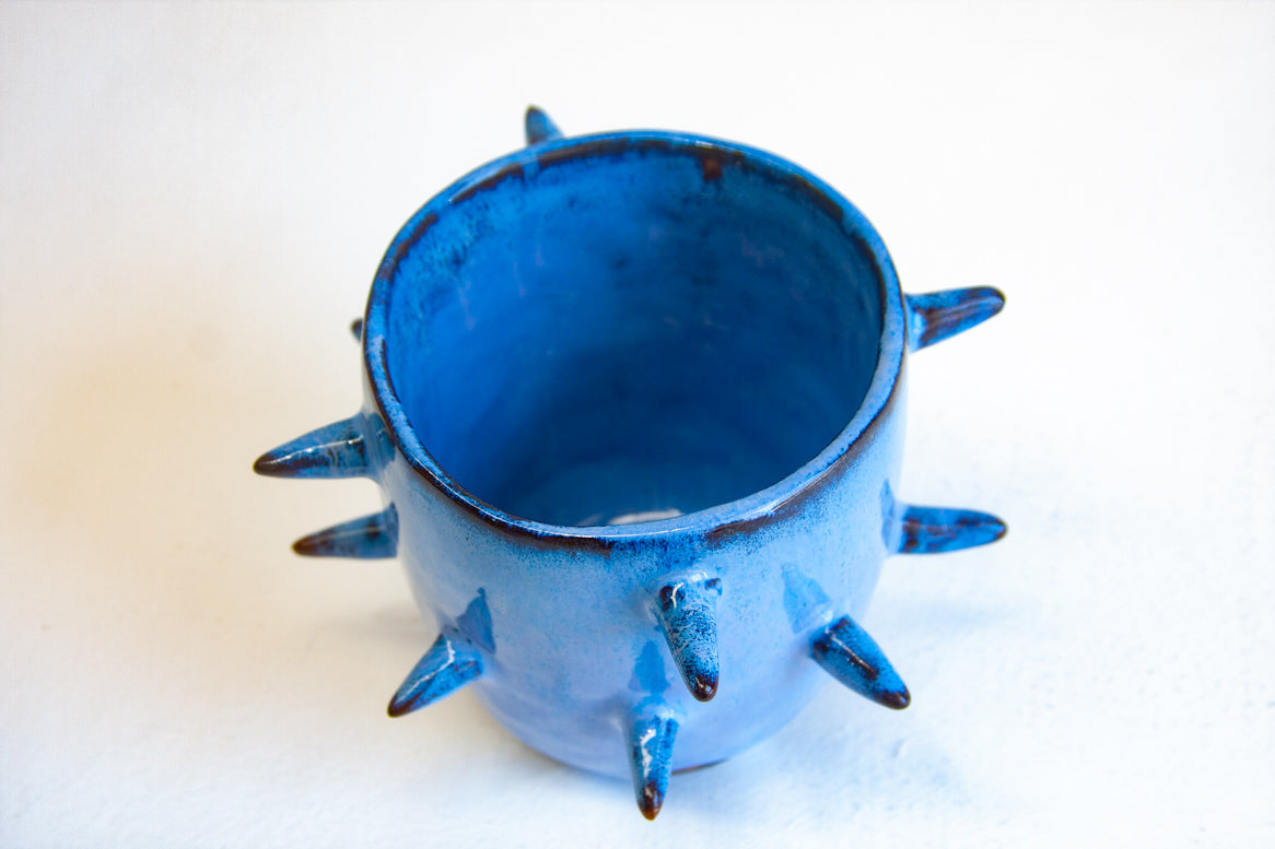 Three-quarter overhead view of empty handmade dark clay spiked vessel; rich cobalt-blue glaze covering the interior and exterior; multiple dark-tipped pointed spikes; dark unglazed clay visible at the base.