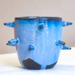 Front-facing side view of empty handmade dark clay spiked vessel; cobalt and periwinkle blue glaze with speckled texture; multiple protruding dark-tipped spikes; prominent unglazed dark clay patches at the base.