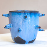 Front-facing side view of empty handmade dark clay spiked vessel; cobalt and periwinkle blue glaze with speckled texture; multiple protruding dark-tipped spikes; prominent unglazed dark clay patches at the base.