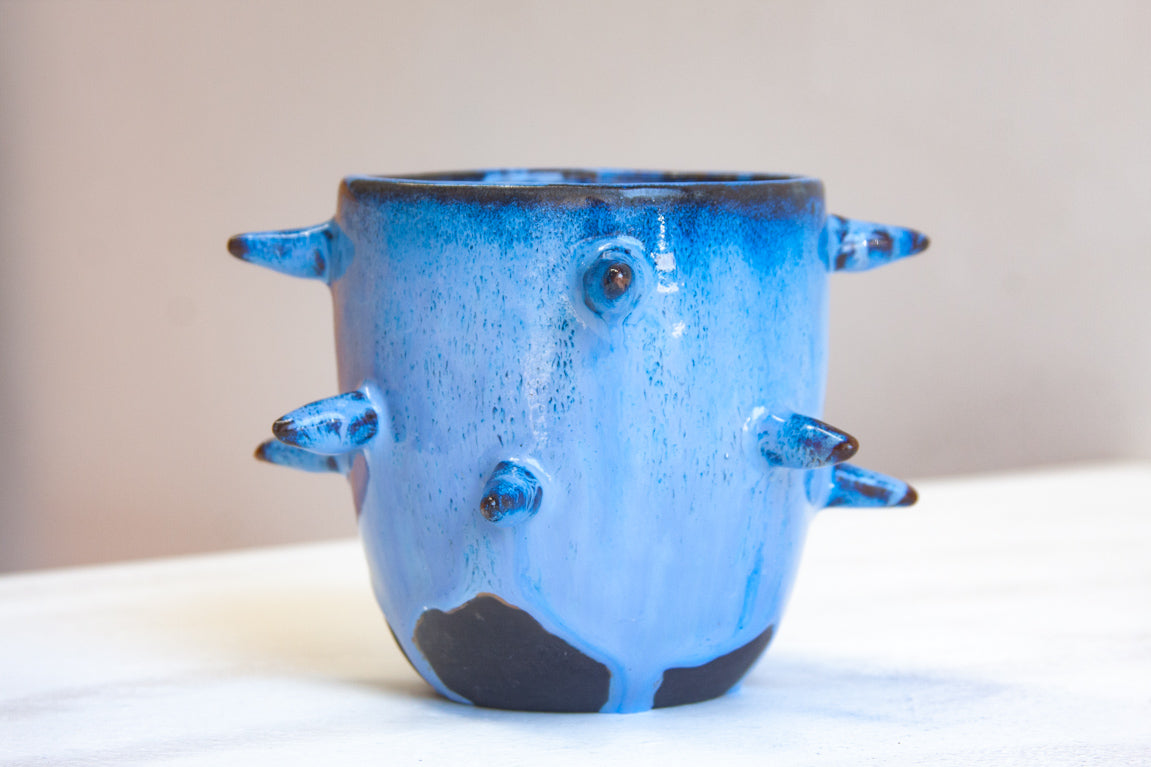 Front-facing side view of empty handmade dark clay spiked vessel; cobalt and periwinkle blue glaze with speckled texture; multiple protruding dark-tipped spikes; prominent unglazed dark clay patches at the base.