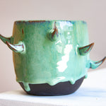 Front-facing side view of empty handmade dark clay spiked vessel on a white plinth against a pink background; bright seafoam-teal glaze dripping to the dark unglazed base; curved brown-tipped clay spikes.