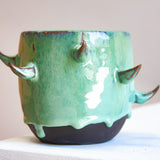 Front-facing side view of empty handmade dark clay spiked vessel on a white plinth against a pink background; bright seafoam-teal glaze dripping to the dark unglazed base; curved brown-tipped clay spikes.