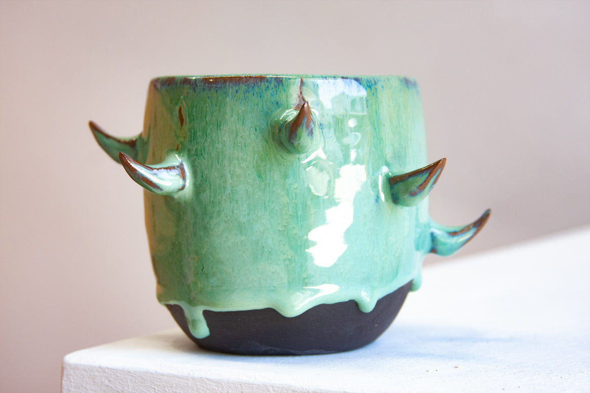 Front-facing side view of empty handmade dark clay spiked vessel on a white plinth against a pink background; bright seafoam-teal glaze dripping to the dark unglazed base; curved brown-tipped clay spikes.