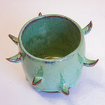 Three-quarter overhead view of empty handmade dark clay spiked vessel; bright seafoam-teal glaze with subtle blue sheen at the rim; curved brown-tipped protruding spikes; dark unglazed clay base.